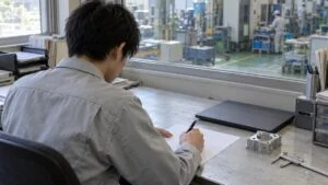 デスクで紙に経歴を書き出す作業着姿の技術者の後ろ姿